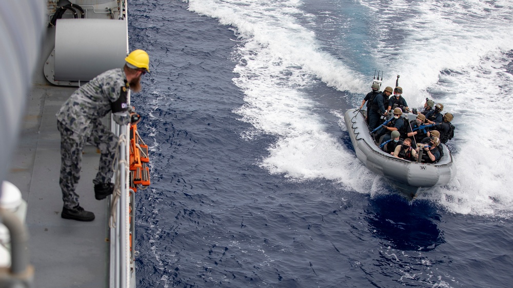 A naval operation showing personnel in a rigid inflatable boat (RIB) approaching a larger warship. A crew member wearing a yellow hard hat and camouflage uniform observes from the ship's deck. The scene appears to depict a maritime boarding team or small boat operation exercise.