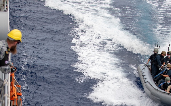 A naval operation showing personnel in a rigid inflatable boat (RIB) approaching a larger warship. A crew member wearing a yellow hard hat and camouflage uniform observes from the ship's deck. The scene appears to depict a maritime boarding team or small boat operation exercise.