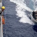 A naval operation showing personnel in a rigid inflatable boat (RIB) approaching a larger warship. A crew member wearing a yellow hard hat and camouflage uniform observes from the ship's deck. The scene appears to depict a maritime boarding team or small boat operation exercise.
