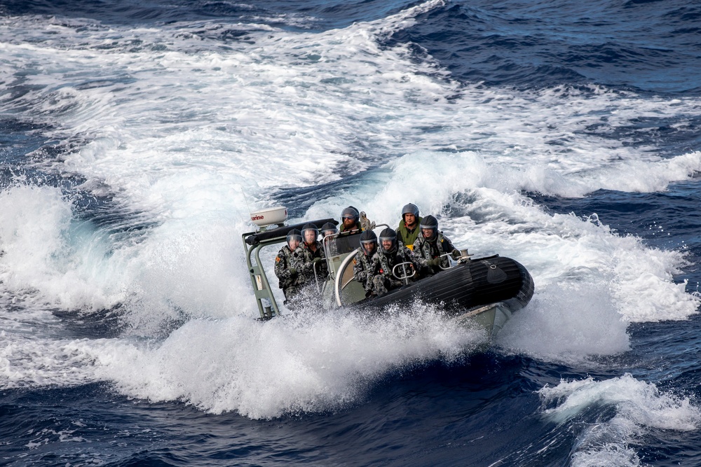 A rigid inflatable boat (RIB) carrying approximately 6-7 military personnel is navigating through rough open ocean waters at high speed. The personnel are wearing camouflage uniforms and helmets with visors, consistent with naval or maritime special operations forces. The boat is generating significant wake and spray as it cuts through choppy waves.