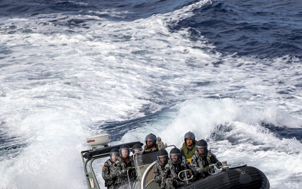 A rigid inflatable boat (RIB) carrying approximately 6-7 military personnel is navigating through rough open ocean waters at high speed. The personnel are wearing camouflage uniforms and helmets with visors, consistent with naval or maritime special operations forces. The boat is generating significant wake and spray as it cuts through choppy waves.