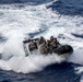 A rigid inflatable boat (RIB) carrying approximately 6-7 military personnel is navigating through rough open ocean waters at high speed. The personnel are wearing camouflage uniforms and helmets with visors, consistent with naval or maritime special operations forces. The boat is generating significant wake and spray as it cuts through choppy waves.