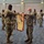 A military ceremony is taking place indoors, likely a casing or uncasing of colors ceremony, where two soldiers in the foreground are handling a unit guidon or flag with yellow and red coloring. Multiple soldiers in U.S. Army OCP (Operational Camouflage Pattern) uniforms are participating in the background, with several pairs also handling flags or staffs. All visible personnel are wearing face masks, suggesting this occurred during the COVID-19 pandemic period.