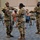 Three U.S. Army soldiers in OCP (Operational Camouflage Pattern) uniforms and face masks are performing what appears to be a military colors or guidon ceremony, likely a casing or uncasing of a unit flag. The ceremony is taking place indoors in what appears to be a conference or community room with civilian observers seated in the background. All three soldiers are actively handling a wooden staff and fabric, suggesting a formal military unit activation or transition ceremony.