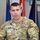 A U.S. Army Colonel named Ellis is speaking at a wooden podium, wearing an OCP (Operational Camouflage Pattern) uniform. His rank insignia (eagle/Colonel) is visible on his chest, along with a U.S. Army tape and name tape. Multiple military and organizational flags are visible in the background.