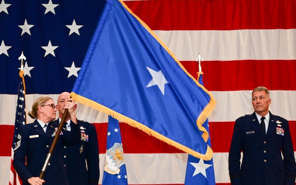 A formal U.S. Air Force military ceremony is taking place on a stage in front of a large American flag backdrop. Three uniformed Air Force personnel are visible, with two handling a blue one-star brigadier general flag while a third officer stands at attention. The presence of multiple one-star flags and the flag-passing action suggests this is a change of command or promotion ceremony.