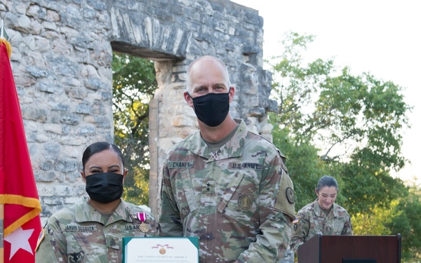 Two U.S. Army soldiers pose together holding a certificate during what appears to be a military awards ceremony conducted outdoors. A taller soldier identified by nametape as 'CHANEY' presents a certificate to a shorter soldier wearing a Purple Heart medal on their uniform. The ceremony takes place near historic stone ruins with military flags and a podium visible in the background.