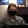 A female military service member wearing U.S. Air Force ABU (Airman Battle Uniform) camouflage and a black face mask is seated at a workstation looking down at a laptop screen. The setting appears to be an indoor office or administrative space. The image was likely taken during the COVID-19 pandemic period based on the mask usage.