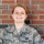 A U.S. Air Force service member in Airman Battle Uniform (ABU) poses for what appears to be an official or semi-official portrait in front of a red brick wall. The name tape reads 'PIANGERELLI' and the uniform displays U.S. Air Force insignia along with what appears to be a Penn State patch. The subject is smiling and has red hair pulled back.