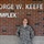 A young female U.S. Air Force service member in ABU (Airman Battle Uniform) stands smiling in front of a brick wall bearing the sign 'George W. Keefe Complex.' She appears relaxed with her hands behind her back, posing for what appears to be an official or commemorative photo. The setting is an exterior wall of a named military or institutional building.