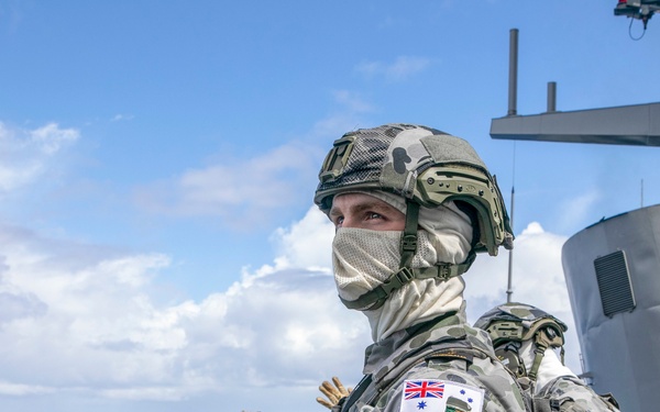 An Australian Navy sailor in full combat gear stands on the deck of a warship at sea. The personnel is equipped with tactical helmet, face covering, body armor, and gloves, and appears to be on active watch or security duty. A second similarly equipped sailor is visible in the background near naval superstructure.