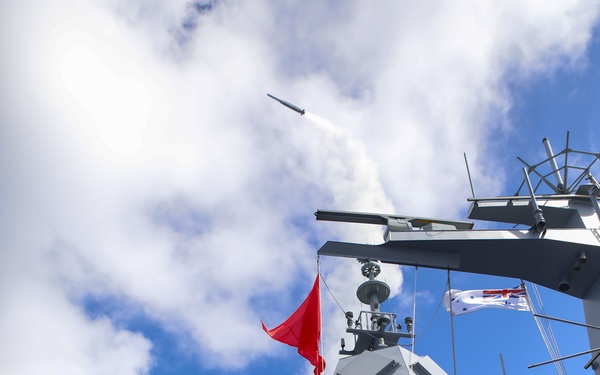 A naval warship is captured mid-missile launch, with a projectile visibly ascending into the sky trailing smoke. The vessel displays a white naval ensign consistent with the Royal Australian Navy, and crew members in protective gear are visible on deck. A red warning/exercise flag is flying from the mast, indicating a live firing exercise is underway.