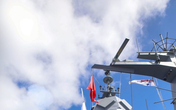 A missile is being launched from a modern naval warship at sea, with a visible smoke trail as the projectile ascends. Personnel in camouflage uniforms are positioned on the deck, and a white naval ensign with a Union Jack (consistent with the Royal Australian Navy White Ensign) is visible flying from the mast. A red signal flag is also displayed on the mast during the firing exercise.