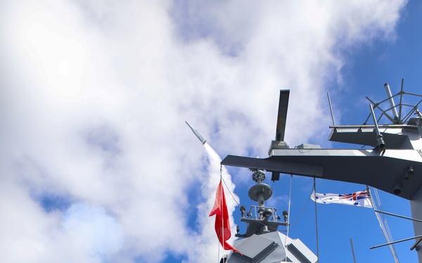 A naval warship is captured mid-missile launch, with a projectile visibly leaving the vessel accompanied by a large plume of white smoke. Armed personnel in camouflage uniforms are positioned on the deck in the foreground. Two flags are visible on the mast structure, including what appears to be a red flag and a naval ensign consistent with the Royal Australian Navy.