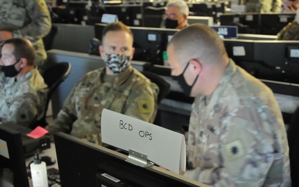 Military personnel in camouflage uniforms and face masks are seated at computer workstations in what appears to be a military operations center or command post. A placard labeled 'BCD OPS' is visible at one of the workstations in the foreground. The setting suggests a training exercise or active operations environment with multiple rows of computer terminals.