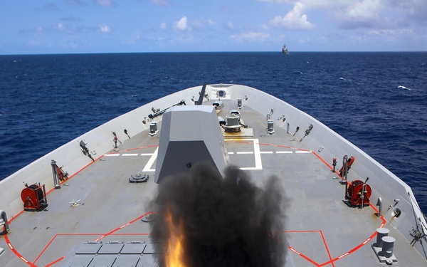 A naval warship is captured mid-firing, with a missile or projectile launching from a Vertical Launch System (VLS) on the bow deck, producing a large plume of black smoke and orange flame. The image is taken from an elevated aft position looking forward along the ship's deck toward the open ocean. A second vessel is faintly visible on the horizon in the background.