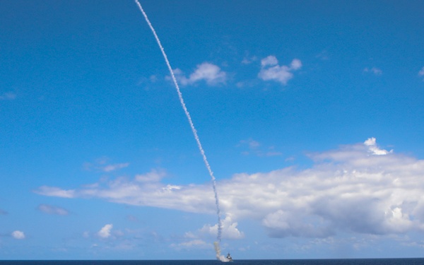 A naval warship is photographed from its bow deck during what appears to be a missile launch exercise at sea. A missile trail is clearly visible ascending steeply into the blue sky, with a smoke plume near the horizon indicating the launch point or a secondary vessel. The image captures an open ocean environment under partly cloudy skies.
