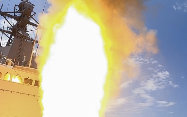 A missile is being launched from the deck of a naval warship at sea, producing an intense white and yellow fireball with billowing smoke. The launch appears to originate from a Vertical Launch System (VLS) mounted on the ship's forward deck. The image was likely captured by an onboard wide-angle camera during a live-fire exercise or combat operation.