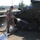 Two military personnel in camouflage uniforms are washing a large armored wheeled vehicle covered in camouflage netting. One soldier is actively spraying the vehicle with a red hose while the other observes. The scene takes place on a wet concrete surface at what appears to be a military base or installation.
