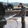 A bearded male soldier in camouflage uniform is using a water hose to wash down what appears to be a military vehicle at an outdoor wash station. The setting appears to be a military base or installation, with other military vehicles and personnel visible in the background. Water is spraying forcefully from the hose nozzle, creating visible water droplets throughout the frame.