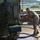 A soldier in camouflage uniform is washing or cleaning a military vehicle using a red hose on a wet concrete surface. The vehicle is covered with camouflage netting and appears to be a large military transport or armored vehicle. The setting appears to be a military base or installation, visible in the background.