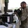 Military personnel in desert camouflage uniforms are conducting an equipment inspection or inventory at what appears to be a covered outdoor staging area. The primary subject, a bearded male service member, is closely examining a black military case or container. Multiple pieces of military equipment and cases are laid out on a work surface.