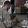 A soldier wearing desert digital camouflage uniform is seated at a workbench, focused on handling or inspecting a piece of equipment or tool. The setting appears to be a covered outdoor or semi-indoor facility, possibly a military base or logistics area. A second person in civilian clothing is visible standing in the background near a large door with a posted sign.