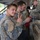 A group of military personnel in camouflage uniforms are gathered at what appears to be a training facility or range. The soldier in the foreground is smiling and making a peace/victory sign gesture toward the camera. Multiple nationalities appear to be represented based on the different uniform patterns visible.