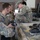 Several military personnel in different camouflage uniforms are gathered around a worn workbench or platform, examining equipment components. At least one soldier appears to wear a Latvian Armed Forces uniform patch ('LATVIJA'), while another wears what appears to be US Army OCP (Operational Camouflage Pattern) uniform. The setting appears to be an indoor military facility or warehouse where equipment inspection or training is taking place.