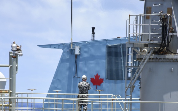 The image shows a large vessel, likely a Canadian naval or coast guard ship, identifiable by the prominent red maple leaf emblem on its blue superstructure. A worker wearing a white hard hat and camouflage clothing stands on the deck amid an extensive network of pipes, railings, and industrial equipment. The scene appears to be taken at sea under partly cloudy skies.