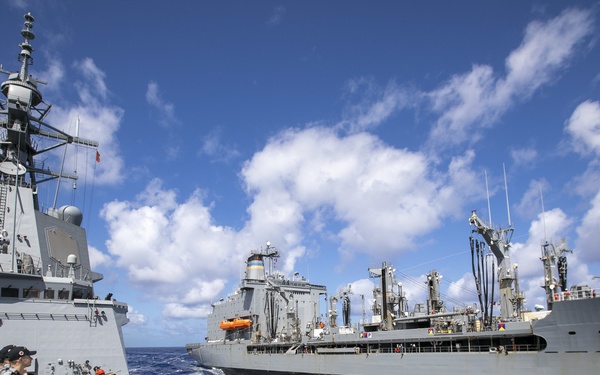The image shows a replenishment-at-sea (RAS) operation between at least two naval vessels underway on open ocean. A warship, likely a destroyer or frigate with visible superstructure, mast, and deck gun, is positioned alongside a larger auxiliary replenishment vessel. Naval personnel are visible on deck managing lines and equipment during the operation.