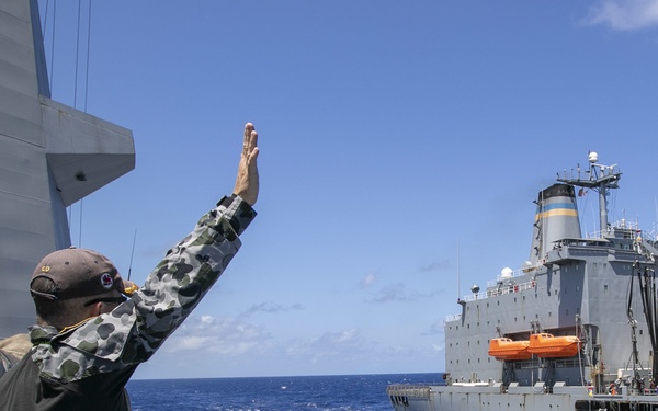 A naval personnel member in Australian Defence Force camouflage uniform raises their hand in a signal or wave toward a large gray replenishment vessel at sea. The scene appears to depict a Replenishment at Sea (RAS) operation, with two ships sailing in close proximity on open blue ocean. The replenishment ship displays a distinctive blue and yellow stripe on its funnel, consistent with a US Military Sealift Command auxiliary vessel.