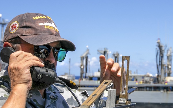 A naval officer in Australian Defence Force camouflage uniform is operating a pelorus (bearing compass) on the bridge wing of a warship while communicating via a handheld radio or sound-powered telephone. The cap insignia reads 'HMAS' followed by partially visible text and 'DDG 39', identifying the vessel as an Australian Navy destroyer. A large grey replenishment or auxiliary vessel is visible in the background, suggesting a replenishment-at-sea (RAS) or close-quarters naval operation.