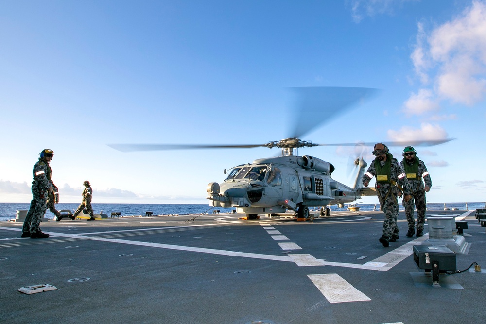 A military helicopter with rotors spinning is positioned on the flight deck of a naval vessel at sea. Several personnel in camouflage uniforms and flight/deck crew gear are moving around the aircraft on the deck. The scene appears to depict either a pre-flight or post-flight operation under clear blue skies.
