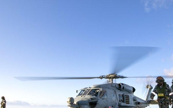 A military helicopter with rotors spinning is positioned on the flight deck of a naval vessel at sea. Several personnel in camouflage uniforms and flight/deck crew gear are moving around the aircraft on the deck. The scene appears to depict either a pre-flight or post-flight operation under clear blue skies.