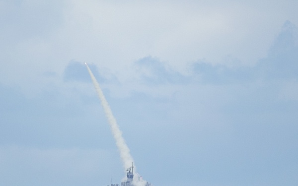 A naval warship is photographed at sea in the act of launching a missile, with a visible smoke trail arcing upward into a partly cloudy sky. The vessel appears to be a modern gray-hulled warship positioned at a distance on open ocean waters. The launch produces a large white smoke plume at the ship's deck level with the missile visible near the top of its trajectory arc.