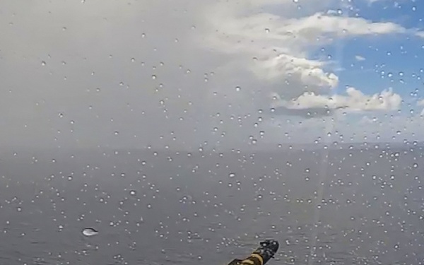 The image captures a missile or rocket in flight over an open ocean, with a bright orange-yellow exhaust plume trailing behind it. The photograph appears to have been taken through a wet glass surface, as water droplets are visible across the entire frame. The sky shows a mix of overcast conditions on the left and clearer skies with cumulus clouds on the right.