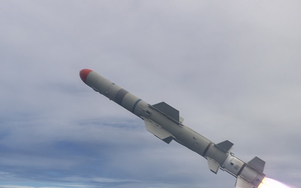 A missile is captured mid-launch from the deck of a naval warship over open ocean. The missile, featuring a red nose cone and active rocket exhaust plume, is ascending at an angle against an overcast sky. The foreground shows deck equipment including what appears to be a gun mount or sensor turret with safety markings visible.