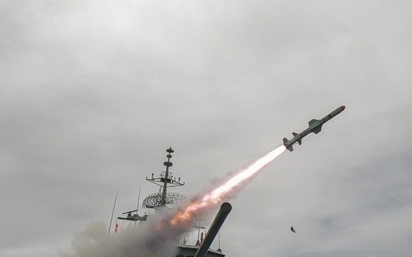 A naval warship is captured in the moment of firing a missile, with a visible rocket trail and exhaust plume rising from the deck. The vessel appears to be a gray military surface combatant equipped with deck guns and superstructure masts. The launch is occurring at sea under a partly cloudy sky.