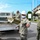 Several U.S. Navy personnel in camouflage uniforms are working around a flatbed utility truck, apparently unloading or handling construction/maintenance materials. The personnel are wearing face masks and hard hats, suggesting safety protocols are in place. The setting appears to be a U.S. military installation, likely in Japan based on the signage and architectural style visible in the background.