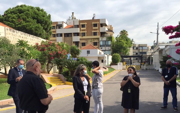 A group of approximately eight to nine people are gathered outdoors in a driveway or courtyard area, appearing to be in a meeting or briefing. Several individuals are wearing face masks, suggesting this was taken during the COVID-19 pandemic period. The setting appears to be a secured compound or institutional facility, with manicured gardens, a security gate, and what appears to be a flag in the upper right corner.