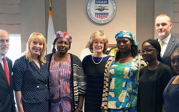 A group of eight individuals pose for a formal photograph in front of a USAID (United States Agency for International Development) seal/emblem. The group appears to be at an official meeting or event, with participants dressed in a mix of Western business attire and traditional African clothing. The setting suggests a professional diplomatic or development-related gathering.
