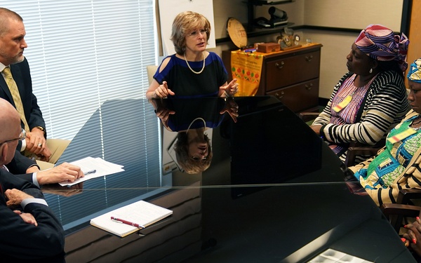 A formal meeting is taking place around a large dark conference table in an office setting. Two groups appear to be engaged in discussion: several individuals in Western business attire on one side, and several women wearing colorful African traditional clothing and head wraps on the other side. Documents and papers are visible on the table, suggesting an official or diplomatic meeting.