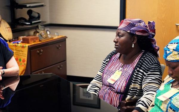 A meeting is taking place around a dark reflective conference table between a woman in Western business attire and two women wearing traditional African clothing and head wraps. The woman on the left appears to be speaking or presenting, while the two women across the table are listening attentively. The setting appears to be a professional office environment.