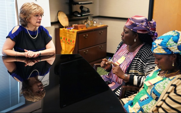 Three women are seated around a reflective black conference table in an office setting, engaged in what appears to be a formal meeting or discussion. One woman on the left is dressed in Western business attire with a pearl necklace, while the two women on the right are wearing traditional African clothing and colorful head wraps. The setting suggests an intercultural or diplomatic meeting context.