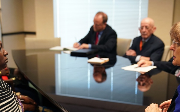 A formal meeting is taking place around a large dark conference table in what appears to be an office or boardroom setting. Several individuals are seated around the table, including a woman wearing a colorful head wrap and striped top on the left, and a woman in a blue dress on the right, appearing to be in direct conversation. Additional participants, including men in business suits, are visible in the background.