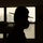 A silhouetted figure is photographed in profile against backlit windows in what appears to be a barracks or institutional interior. The person is wearing a wide-brimmed campaign/drill sergeant hat and a face mask, rendered entirely in dark silhouette. The high-contrast lighting creates a dramatic, anonymous composition.