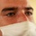 A close-up portrait of a young adult male wearing a white surgical/medical face mask. The subject has green-hazel eyes, dark short hair, dark eyebrows, and visible freckles across the nose and cheeks. The background is a soft, blurred yellow-cream color.