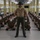 A U.S. Marine Corps drill instructor stands at attention in the center aisle of a military barracks, facing the camera with hands clasped behind his back. Behind him, two rows of recruits in camouflage uniforms sit cross-legged on the floor in a disciplined formation. All individuals are wearing face masks, consistent with COVID-19 health protocols during military training.