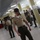The image shows a U.S. Marine Corps drill instructor in a barracks bay, wearing the distinctive campaign cover (drill instructor hat) and tan uniform with ribbons. Multiple personnel in camouflage uniforms are present in the background, and all visible individuals are wearing face masks, suggesting this was taken during the COVID-19 pandemic period. The setting appears to be a recruit training barracks with bunk beds visible along the walls.